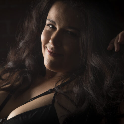 Flirty boudoir photo of a woman sitting up against a sofa, backlit by late afternoon light. She's wearing a strappy black teddy, and one hand is up to her hair as she looks over, smiling sweetly at the camera
