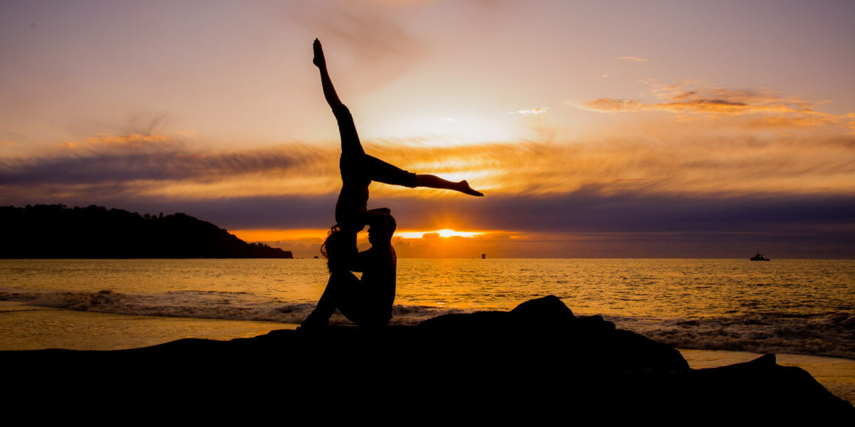 Nude couple in silhouette doing acro yoga together on rocks at the beach during sunset.