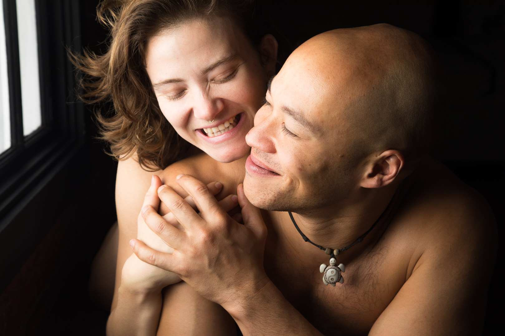 Couples boudoir image with a woman and a man, both nude and smiling, holding hands as the woman embraces the man from behind.