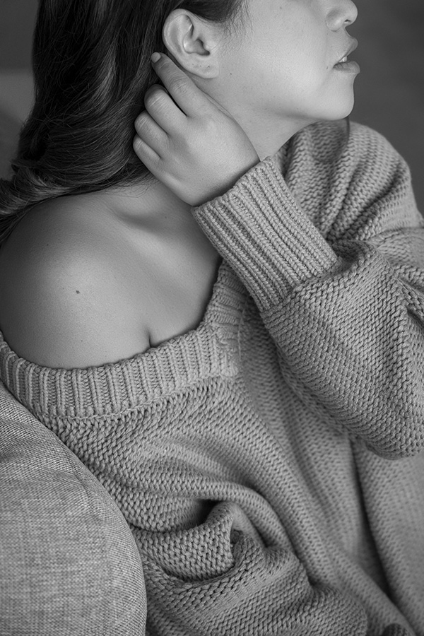 Delicate boudoir image of a woman in a knit sweater off her shoulder. She's looking off to the side as her hand is playing with her hair.